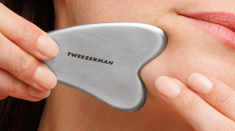 Person using a silver Gua Sha tool on their neck with 'Tweezerman' branding.